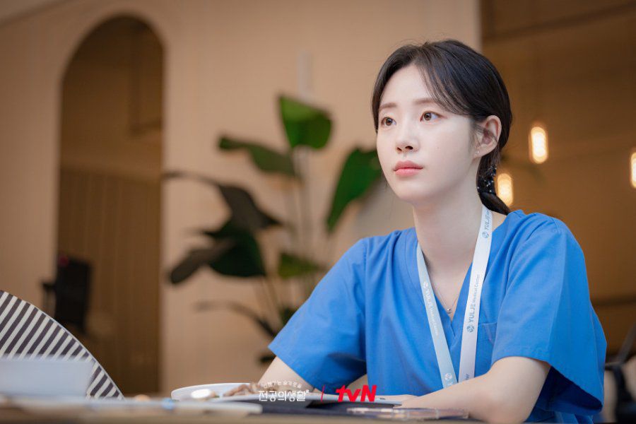 Karakter Resident Playbook, Pyo Nam-kyung, menggunakan scrub biru ala dokter, sedang menatap dalam lawan bicaranya di kafe.